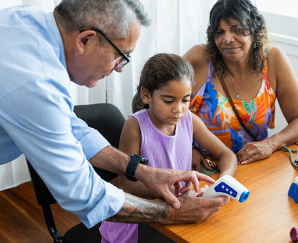 Middle aged Pacific Islander doctor shows young Aboriginal patient her temperature on a digital thermometer, while her Grandmother looks on in support