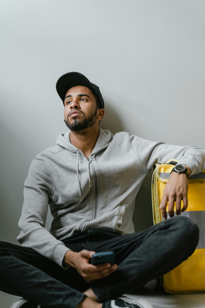 A young man wearing hat leaning against the wall holding a phone with a look of contemplation on his face
